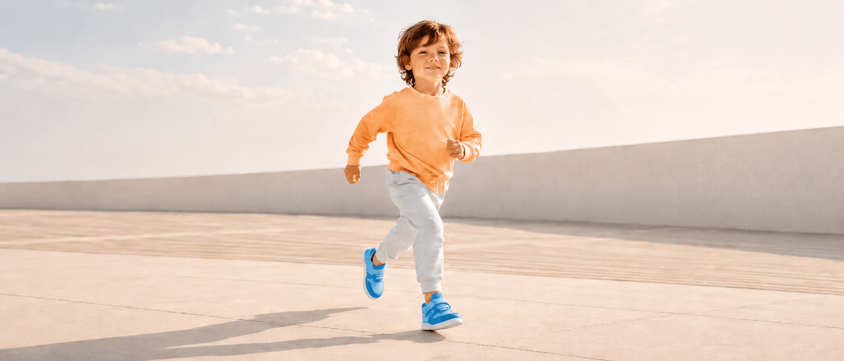 Young boy running outdoors smiling, wearing an orange sweater and blue barefoot sneakers from Pas Naturels, front view.