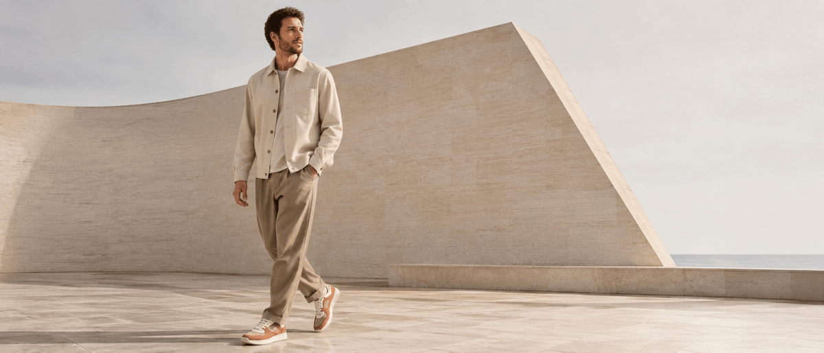 Man walking outdoors in a beige overshirt and trousers, wearing tan and white barefoot sneakers from Pas Naturels.