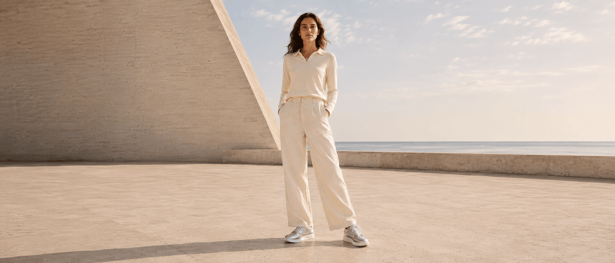 Woman standing outdoors by the sea in a cream polo top and trousers, wearing silver metallic barefoot sneakers from Pas Naturels.